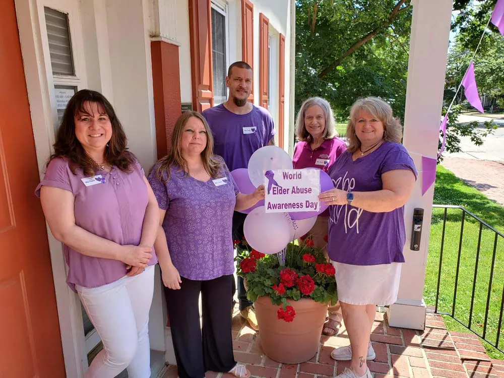 Group of women wearing purple holding up sign posing for photo outside on porch