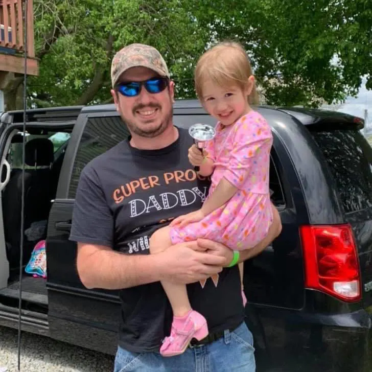 Little girl being held up in dad's arms outside in front of car